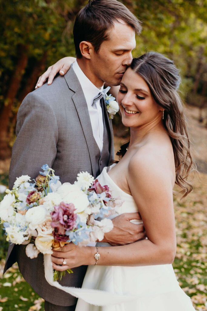 Groom kissing brides forehead while bride holds bridal bouquet.